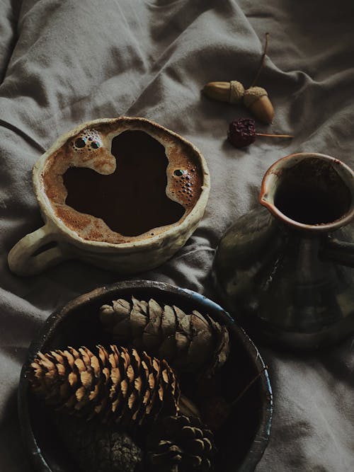 A Cup of Drink and a Bowl of Conifer Cones on Gray Fabric