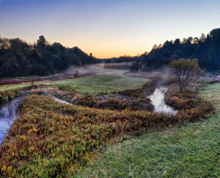 Serene landscape in Misha Mokwa, Wisconsin with misty fields and a curving stream at sunrise.
