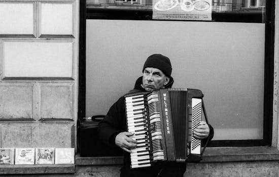 A street performer plays the accordion outside a building in a monochrome setting. CDs displayed nearby.
