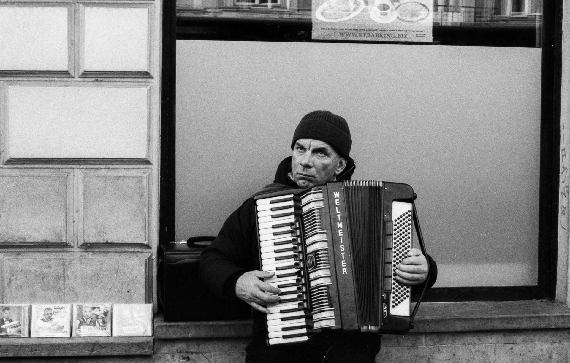 A street performer plays the accordion outside a building in a monochrome setting. CDs displayed nearby.