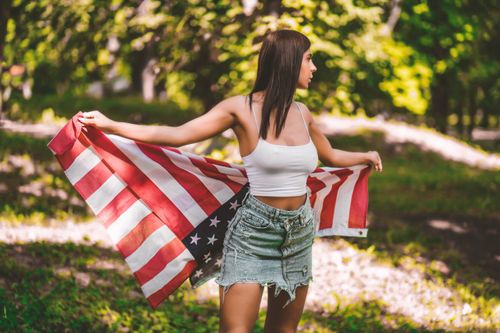 Free Young woman in crop top and denim skirt holding an American flag in a sunny forest. Stock Photo