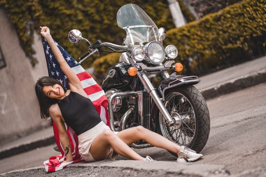 Smiling woman with American flag posing by a motorcycle on an outdoor street.