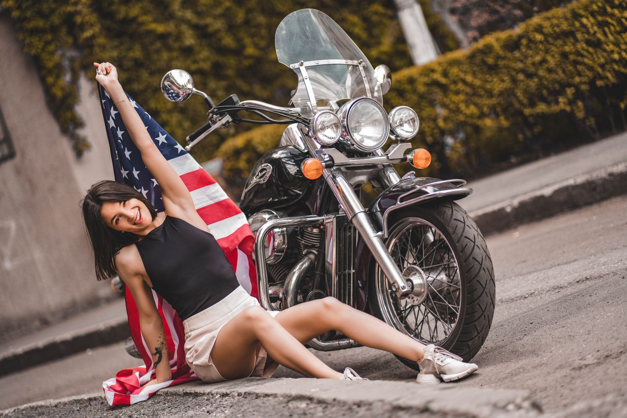 smiling woman with american flag posing by a motorcycle on an outdoor