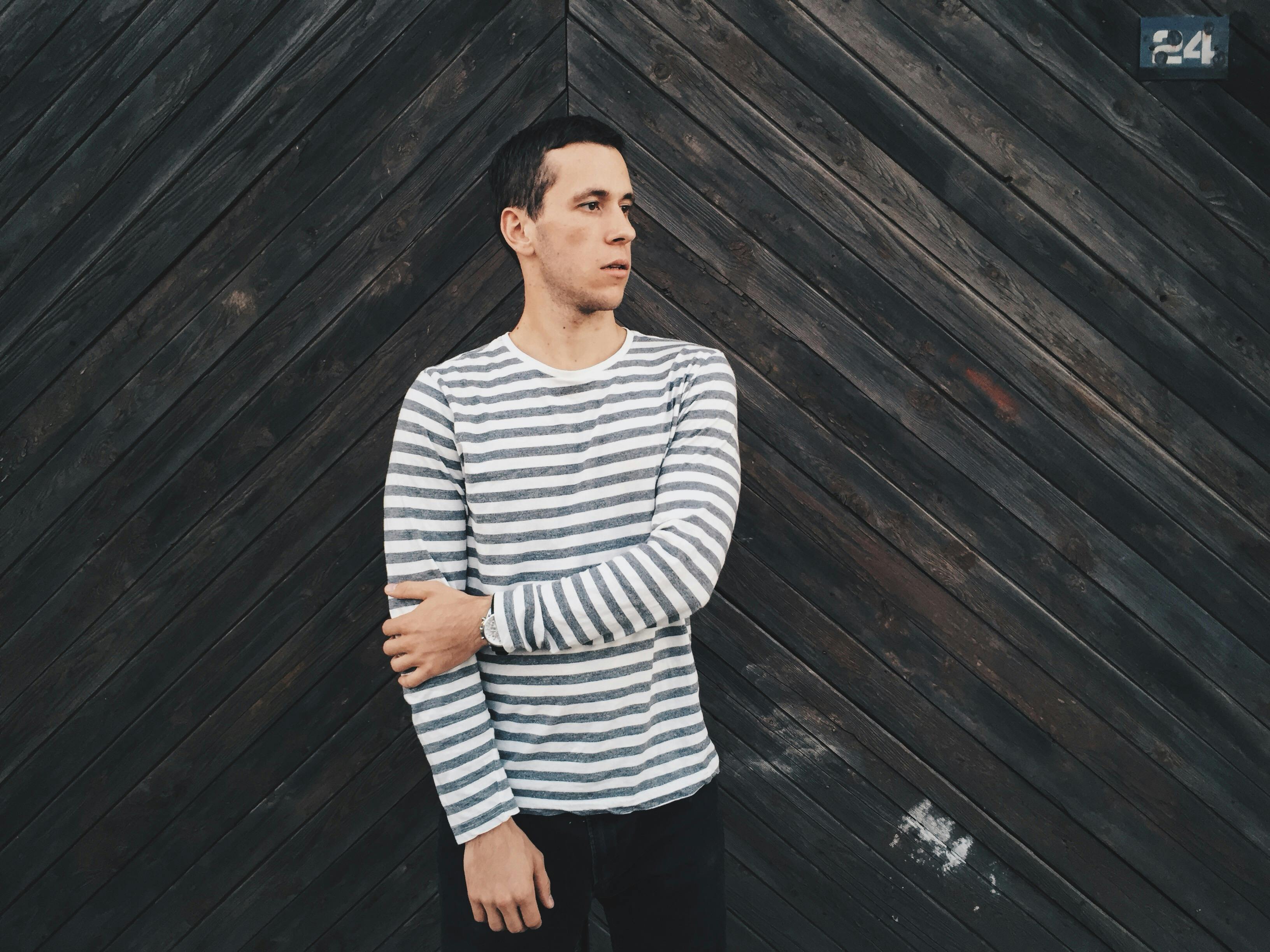 A man wearing a striped shirt stands against a textured wooden wall looking away.