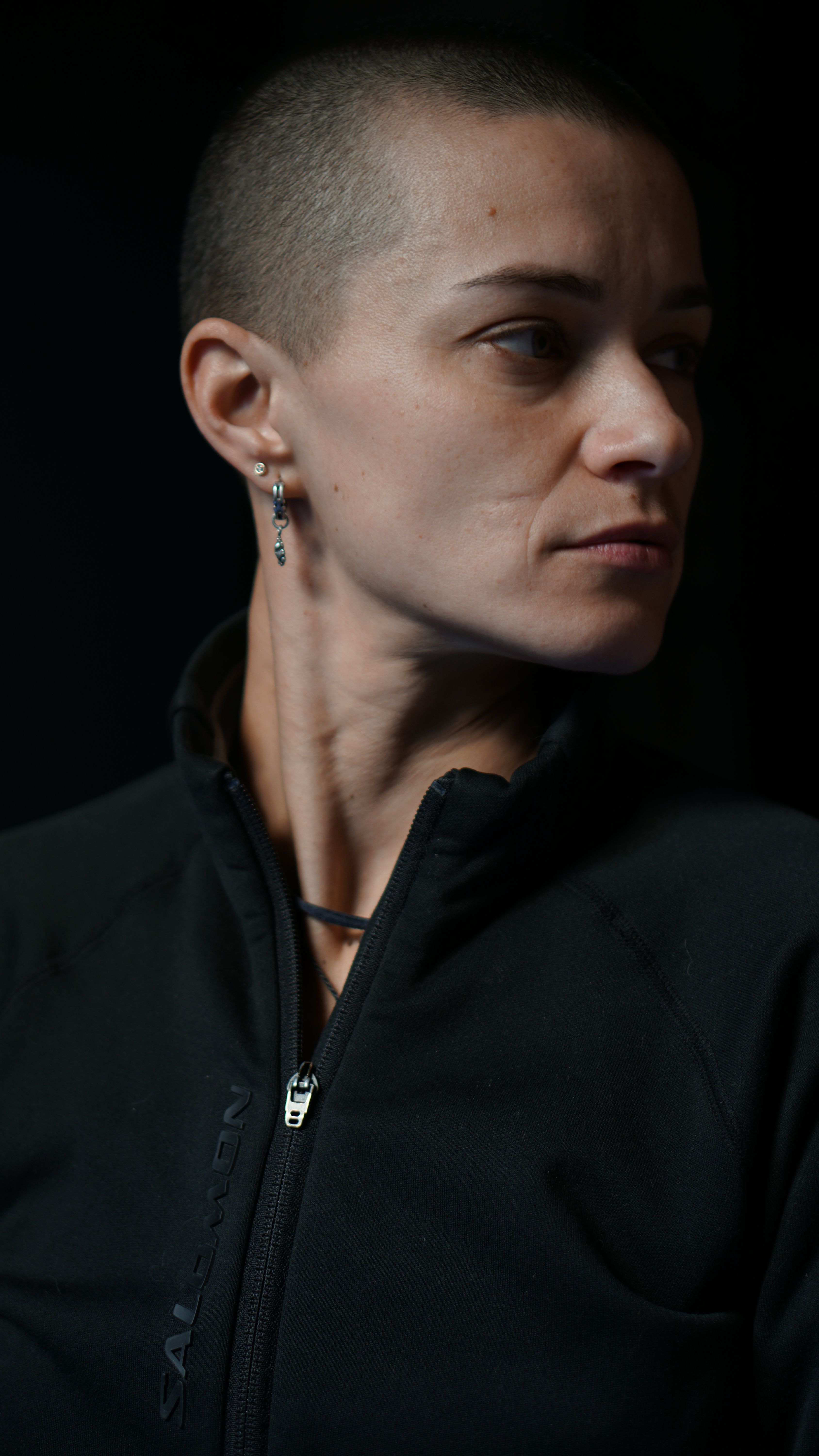 Close-up portrait of a woman with a shaved head wearing a black jacket, thoughtful expression.