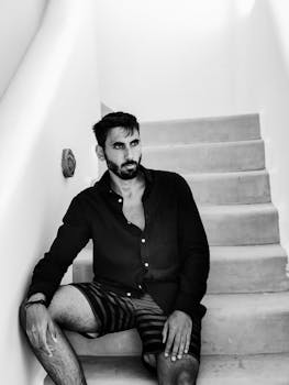 Black-and-white portrait of a fashionable man sitting on stairs in Mykonos, exuding a cool and relaxed vibe.
