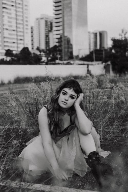 Grayscale Photo of a Young Woman in Spaghetti Strap Dress Sitting on Grass Field