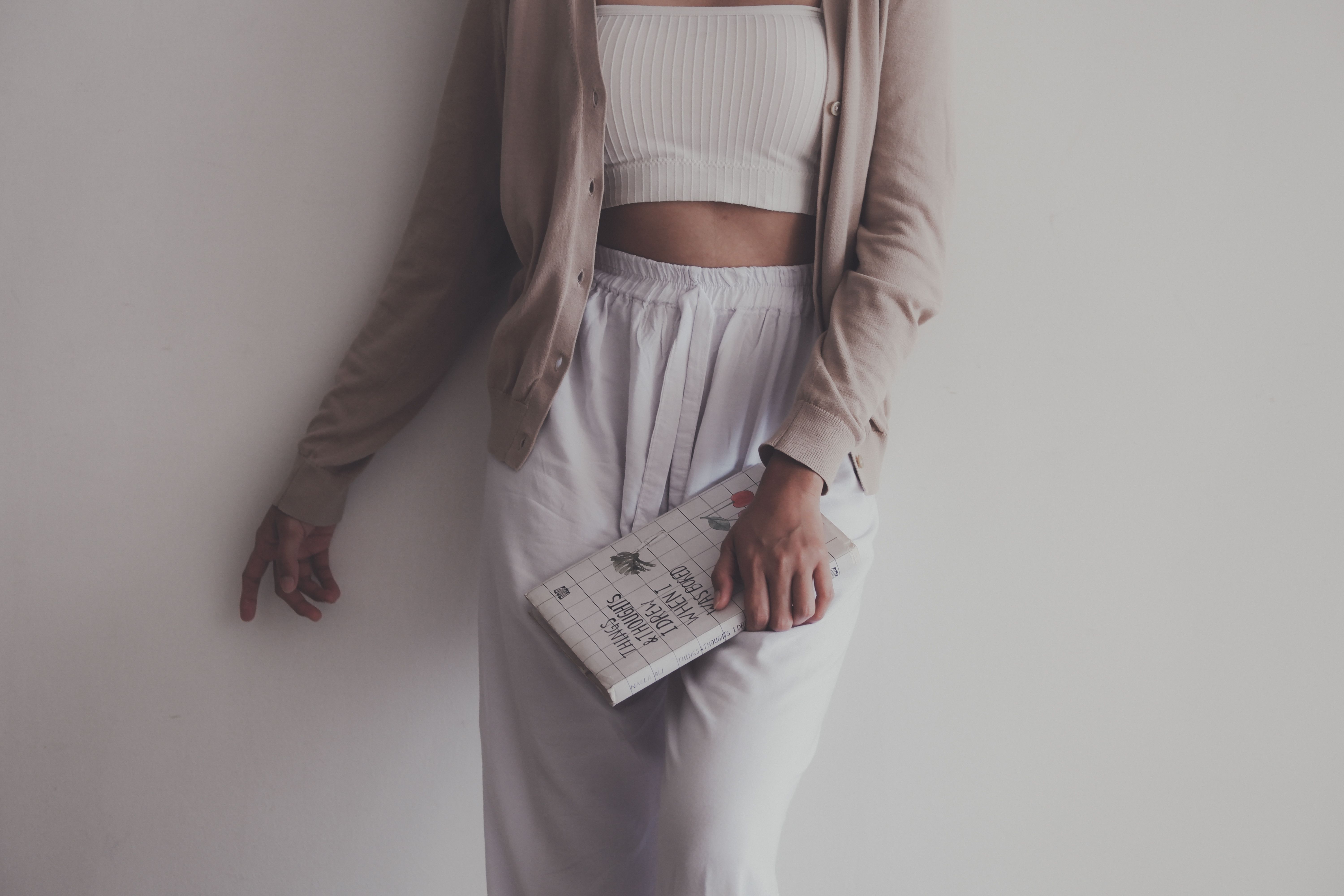 A person in a beige cardigan and white outfit holding a book against a white wall.