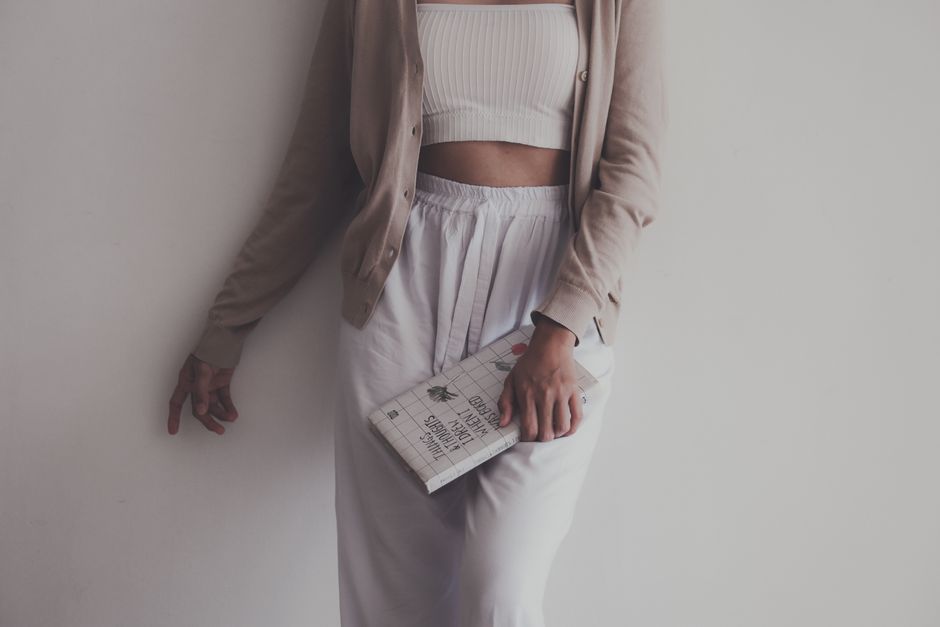 A person in a beige cardigan and white outfit holding a book against a white wall.
