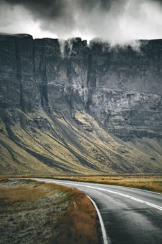 A winding asphalt road leads towards dramatic cliffs under a moody, overcast sky in Iceland, showcasing natural beauty.