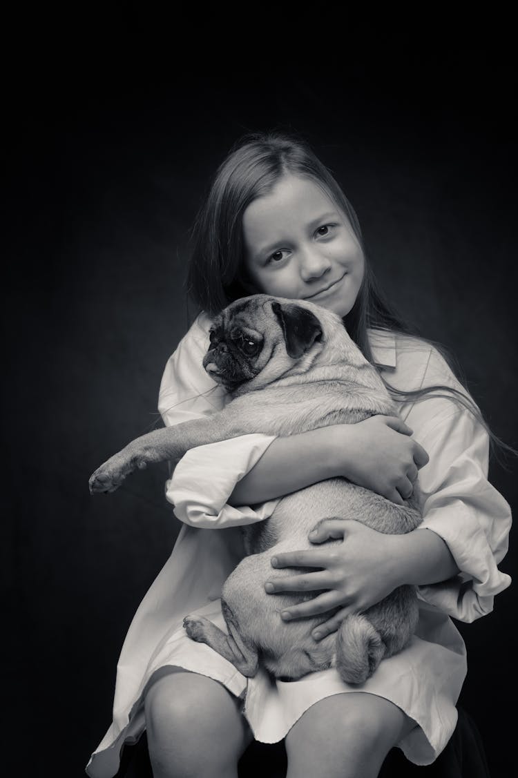 Grayscale Photo Of Young Girl Sitting And Embracing A Pug Dog