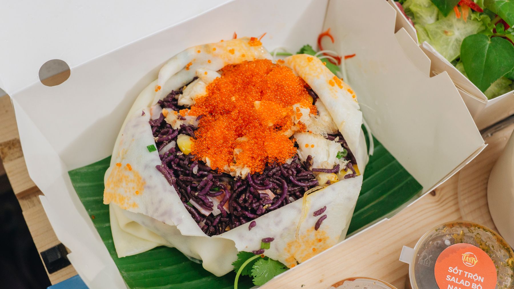 A vibrant Asian fusion salad wrap topped with orange roe and presented in a takeaway box.