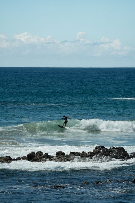 Lone surfer skillfully riding ocean waves with rocky shoreline foreground under a clear sky.