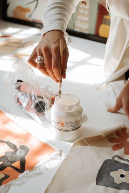 A hand dipping a brush into paint while creating art on fabric in a sunlit studio.