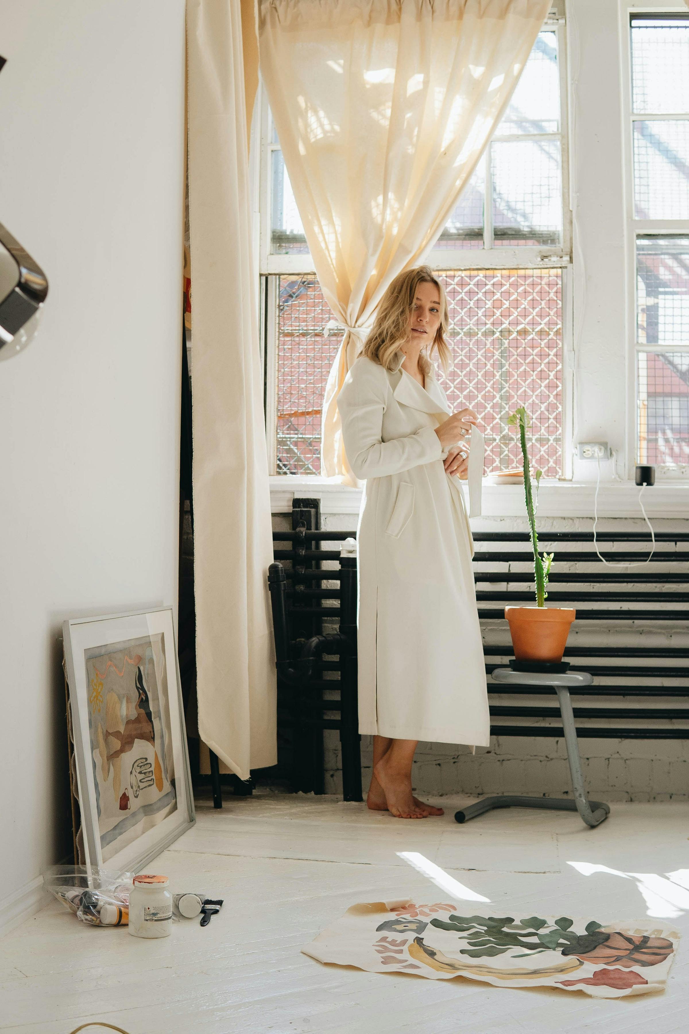 Woman in a white coat standing in a bright, minimalist room with large windows.