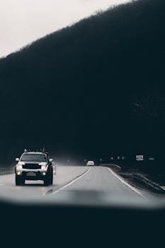 A lone SUV navigates a wet road through a misty mountain landscape, emphasizing travel and adventure.
