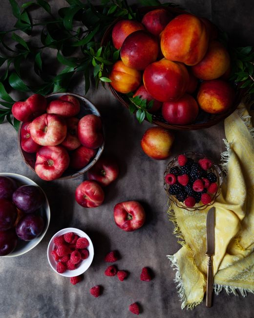 A colorful, fresh assortment of summer fruits including nectarines, plums, and berries on a rustic tabletop with leafy greens.