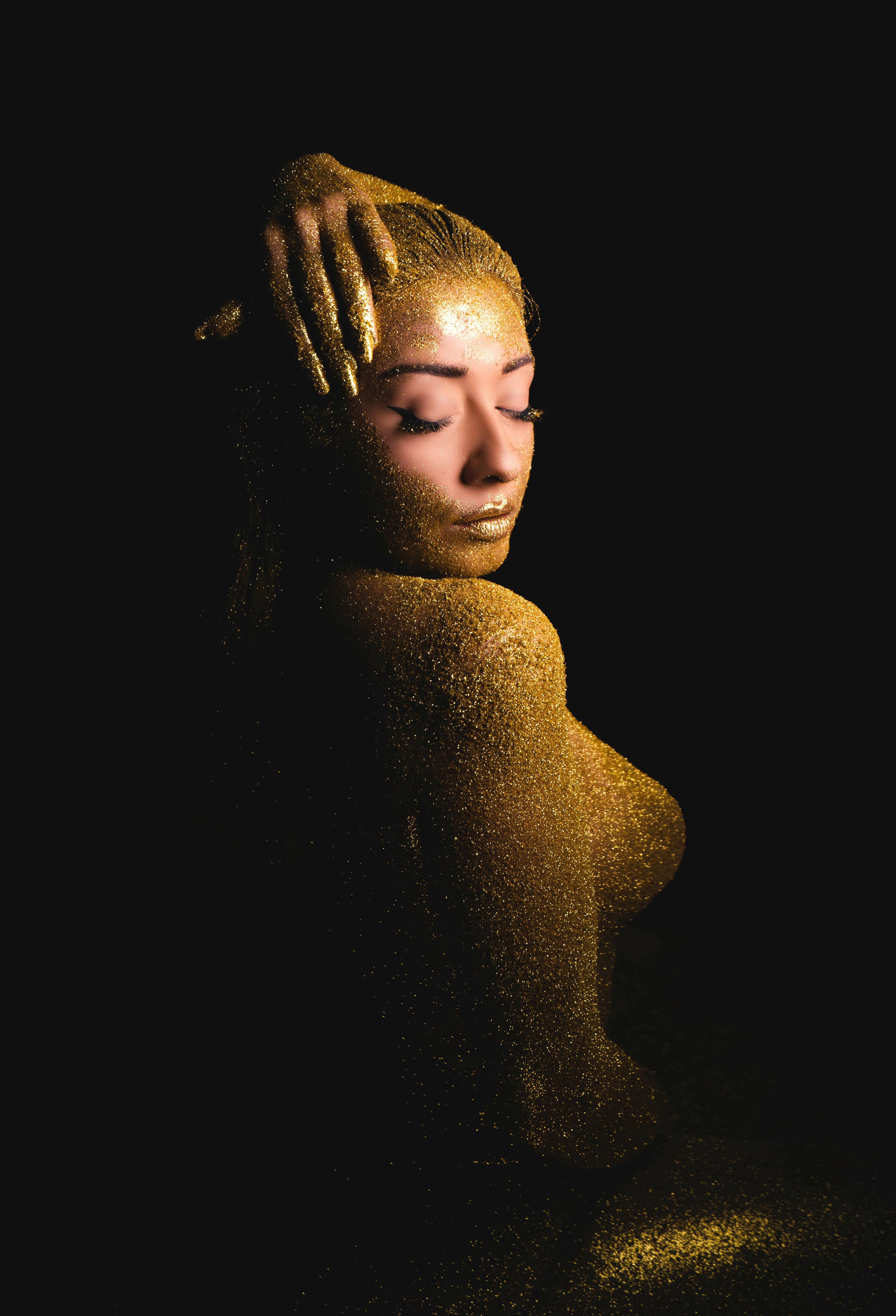 Artistic portrait of a woman covered in golden glitter against a dark background.