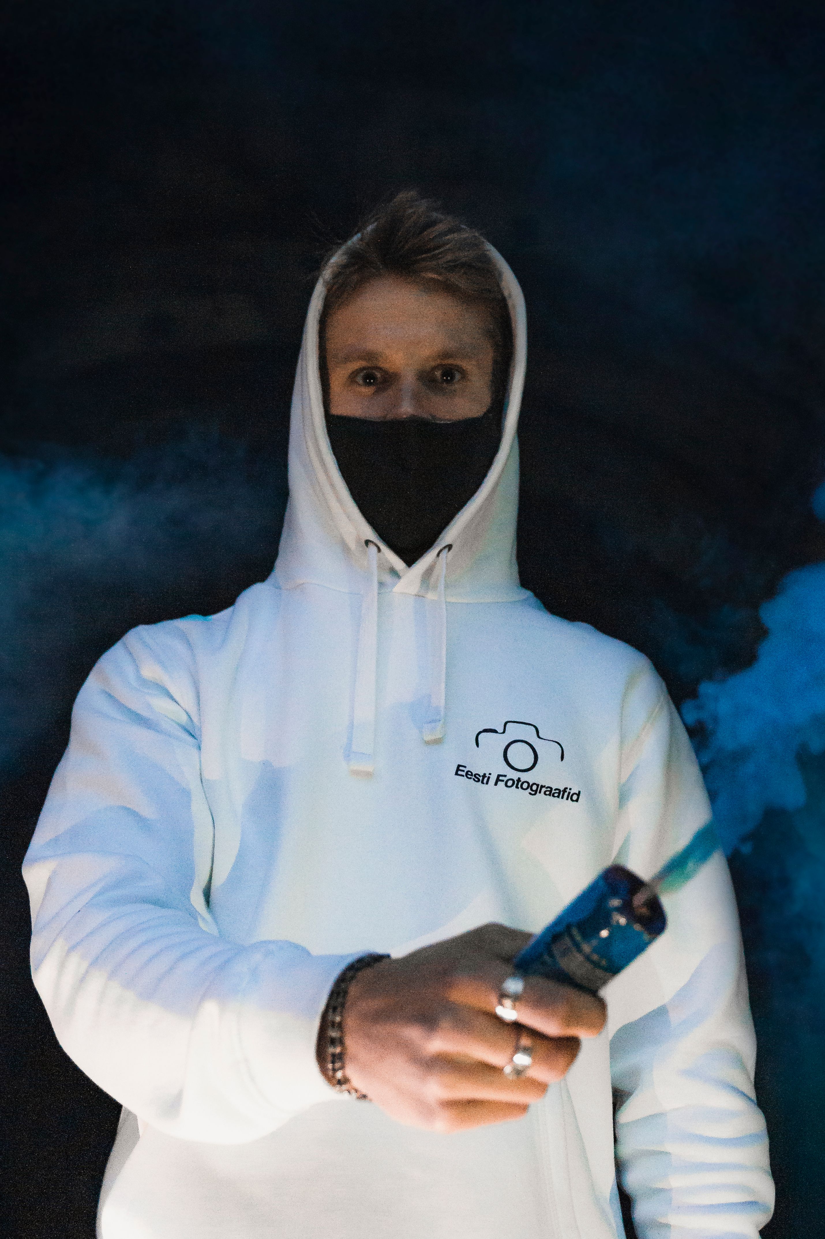 Portrait of a masked man in white hoodie with colored smoke against a dark background.