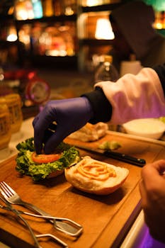 A chef wearing gloves prepares a fresh gourmet burger with lettuce and tomato in a vibrant restaurant setting.