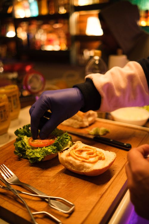 Free A chef wearing gloves prepares a fresh gourmet burger with lettuce and tomato in a vibrant restaurant setting. Stock Photo