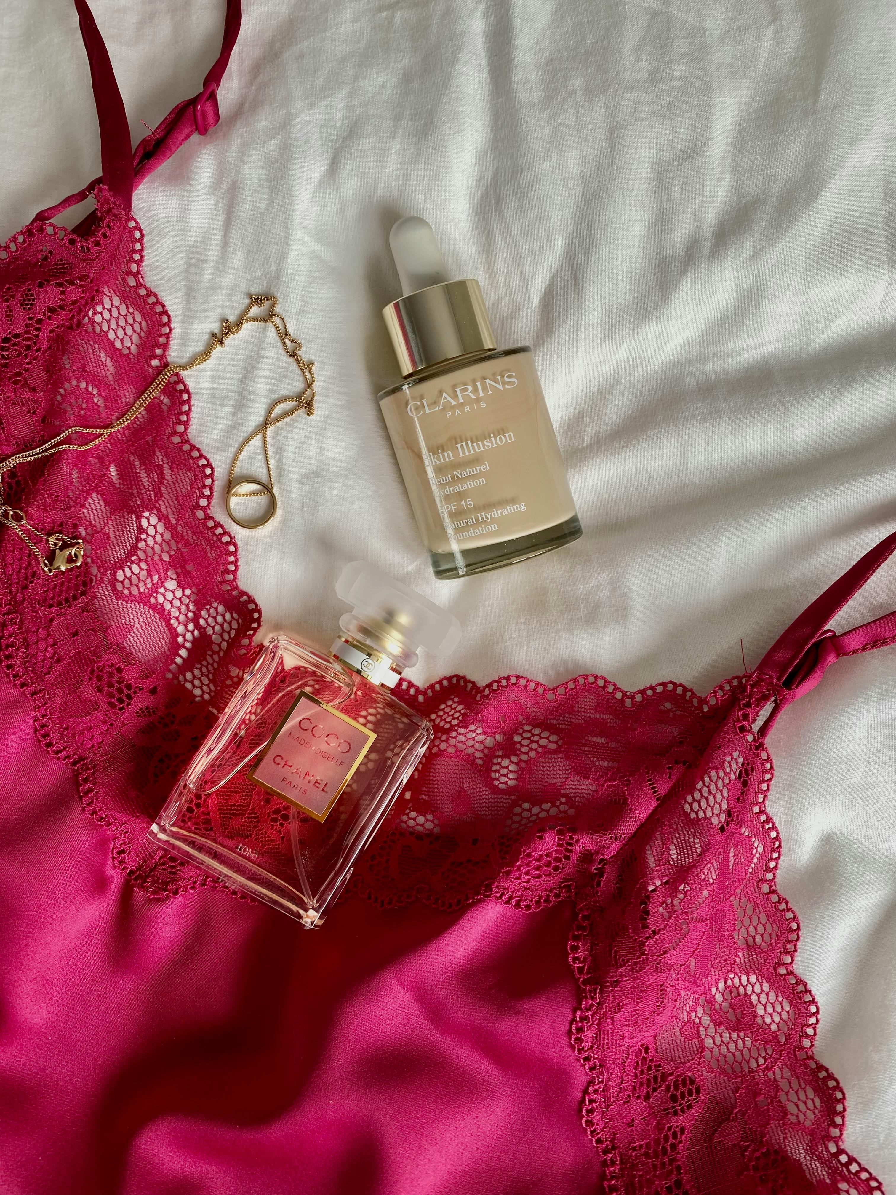 Stylish flat lay of pink lingerie, perfume, and beauty serum on white fabric.