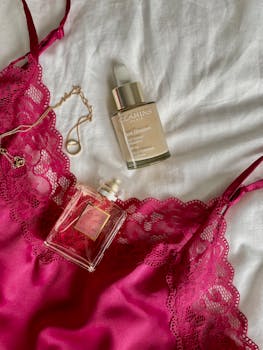 Stylish flat lay of pink lingerie, perfume, and beauty serum on white fabric.