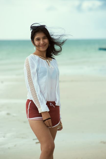 Asian woman enjoying a windy beach day wearing a white top and red shorts, smiling with the sea in the background.