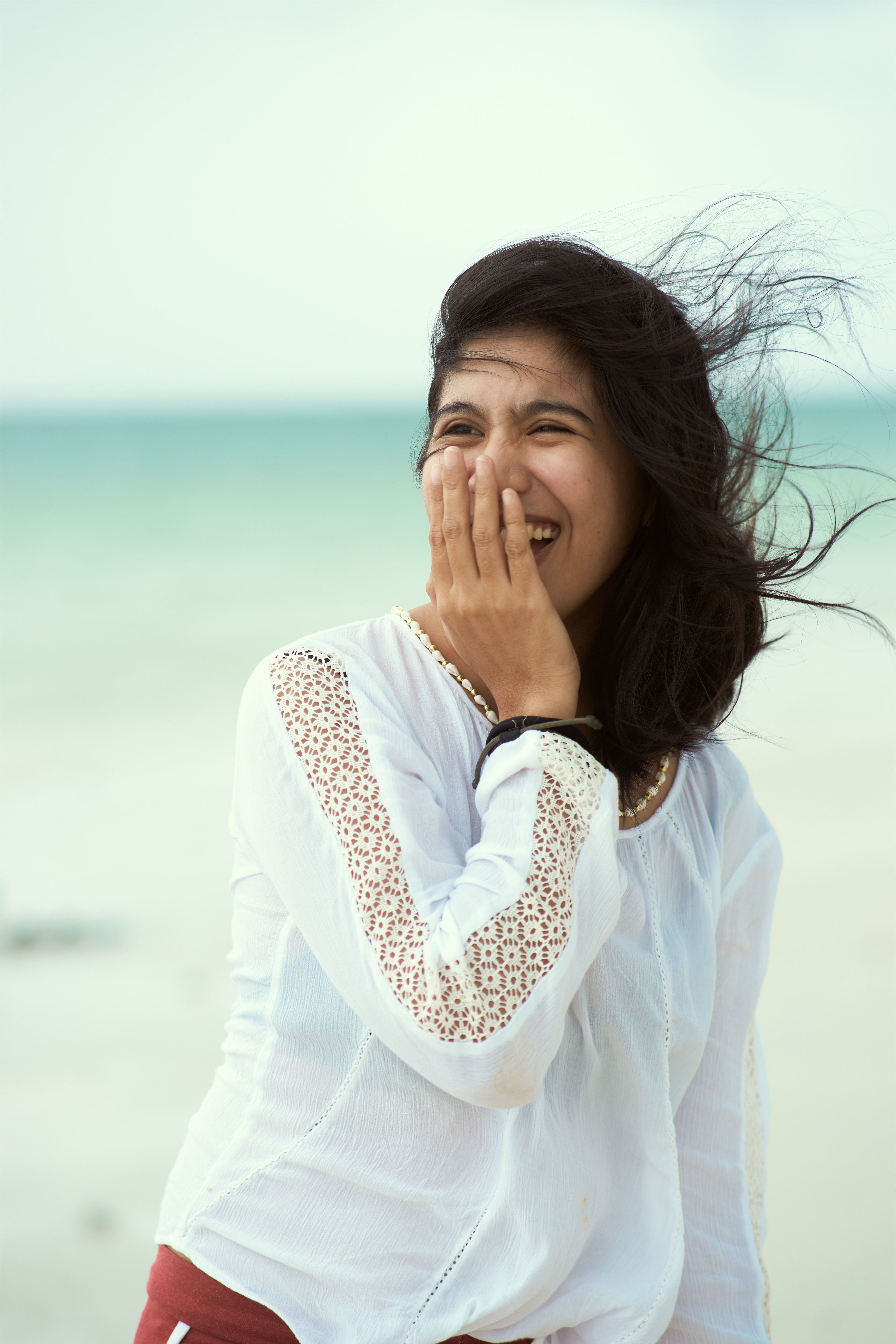 A joyful woman in a white top smiles with the beach and wind in the background.