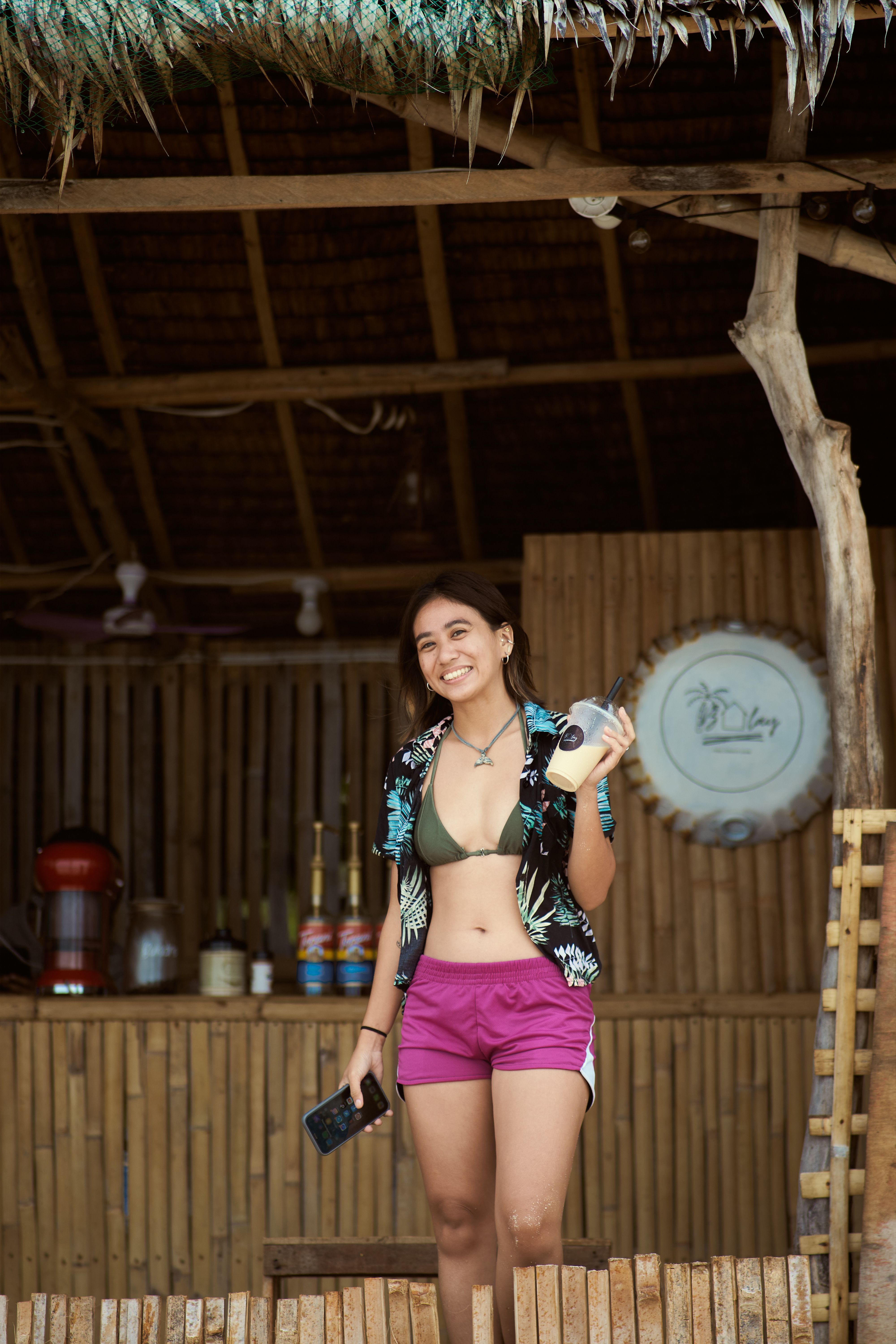 Smiling woman in summer clothing at a rustic bamboo bar, radiating positive vibes.
