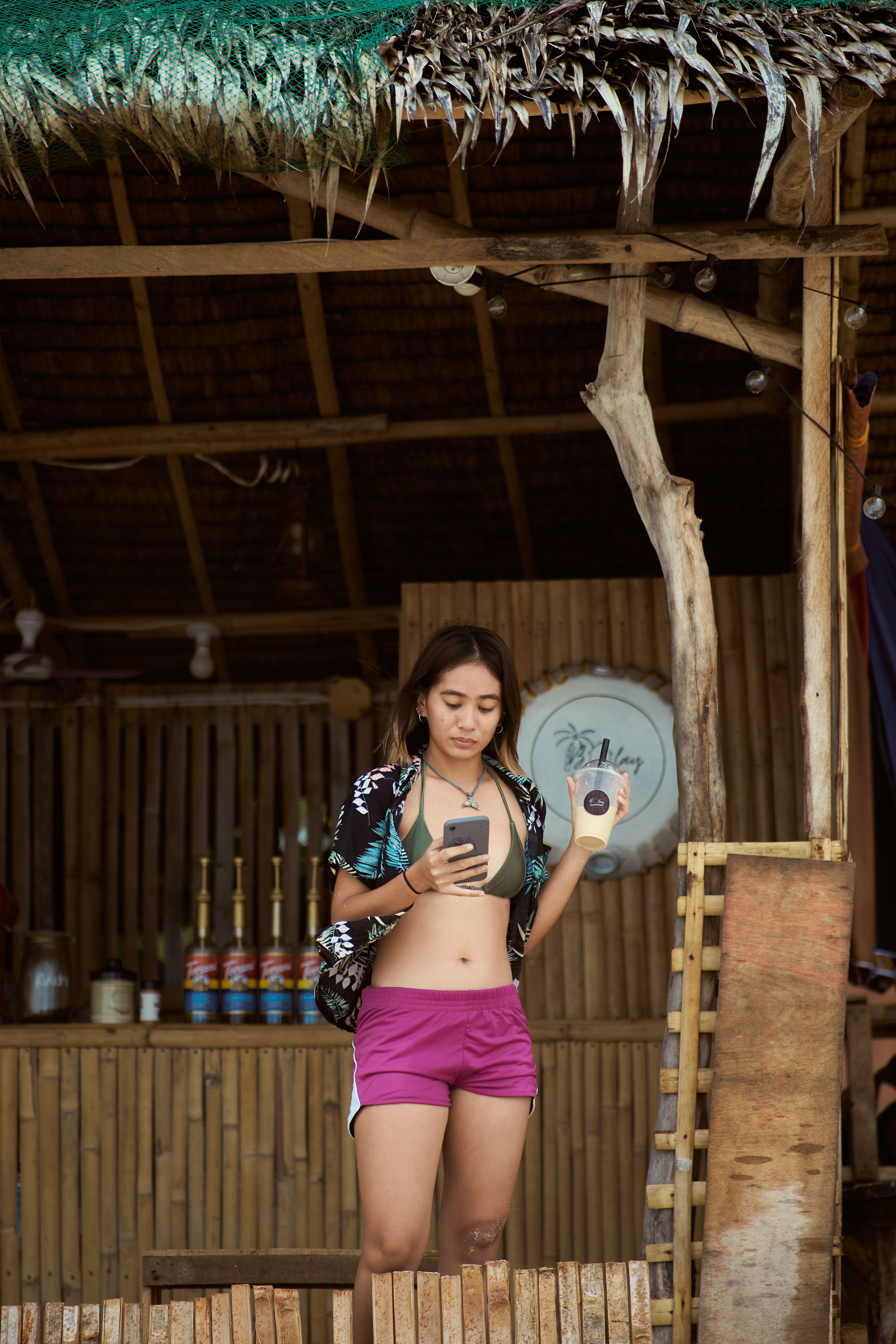 Young woman in summer attire holding milk tea and smartphone at a bamboo beach hut.