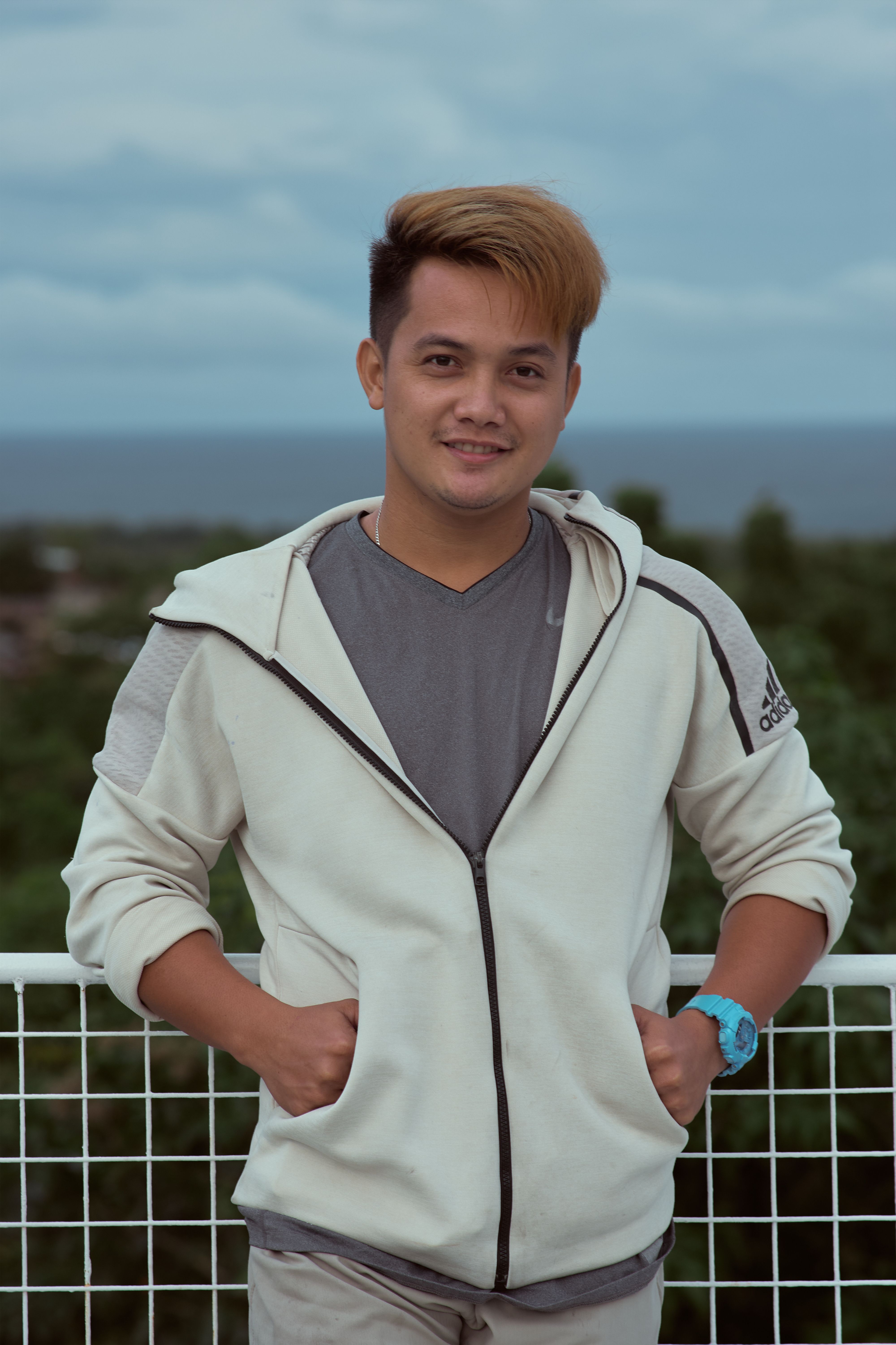 Young man smiling outdoors with a blue sky and greenery in the background, wearing a white hoodie.