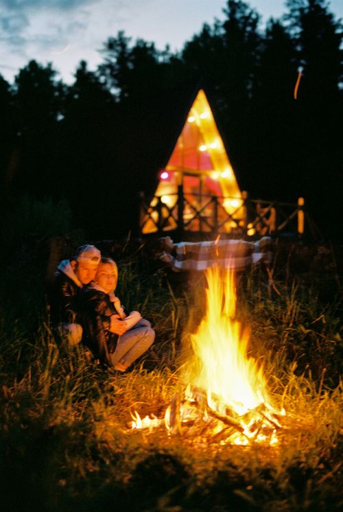 Free A young couple enjoys a campfire outside an illuminated cabin at night in the forest. Stock Photo