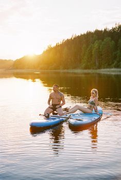 A couple sits on paddleboards on a serene lake at sunset, enjoying nature.