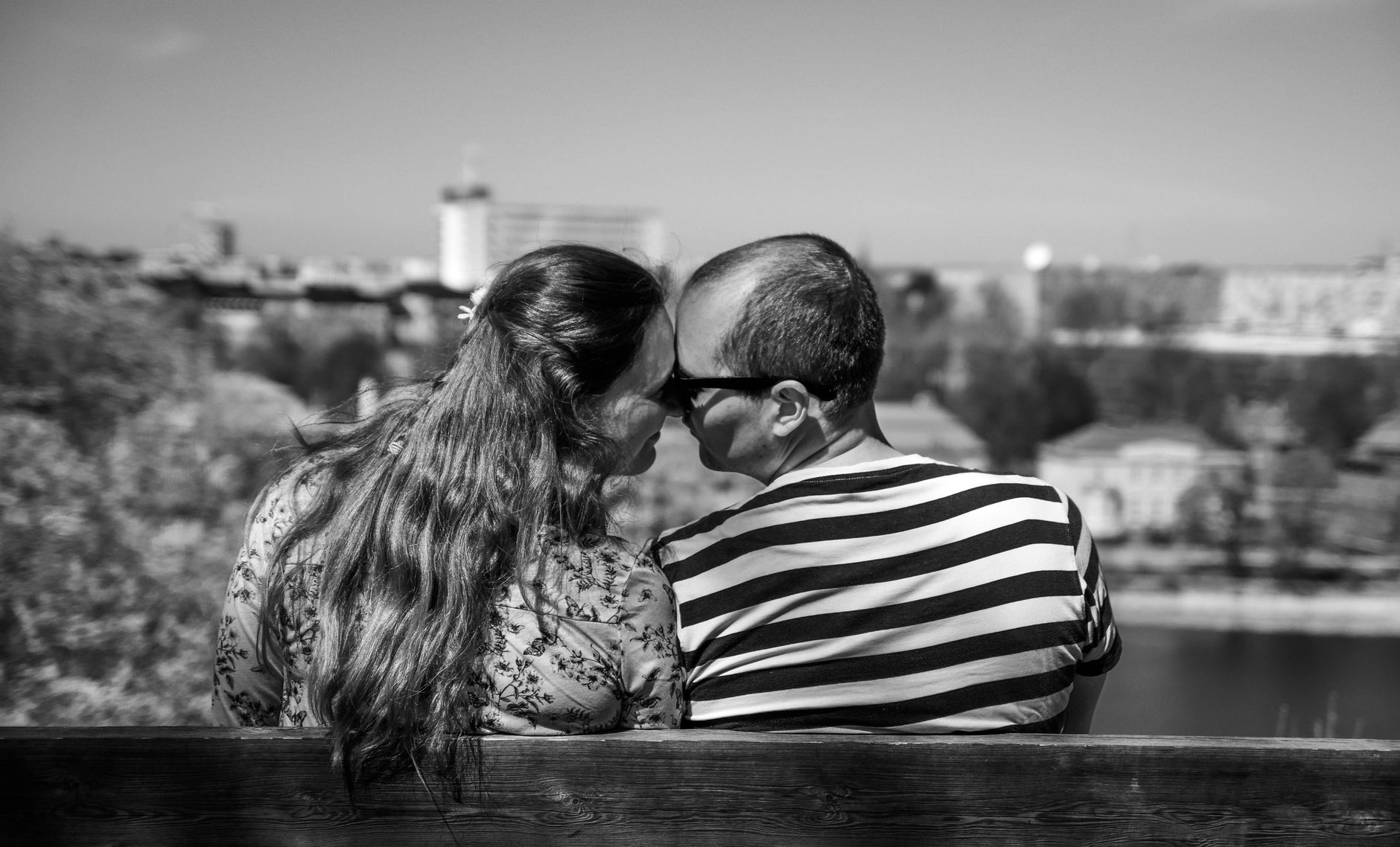 Black and white photo of a couple sharing a quiet, close moment on a bench overlooking a city.