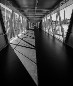 Black and white photo of a sleek indoor urban walkway with steel framework and natural lighting.