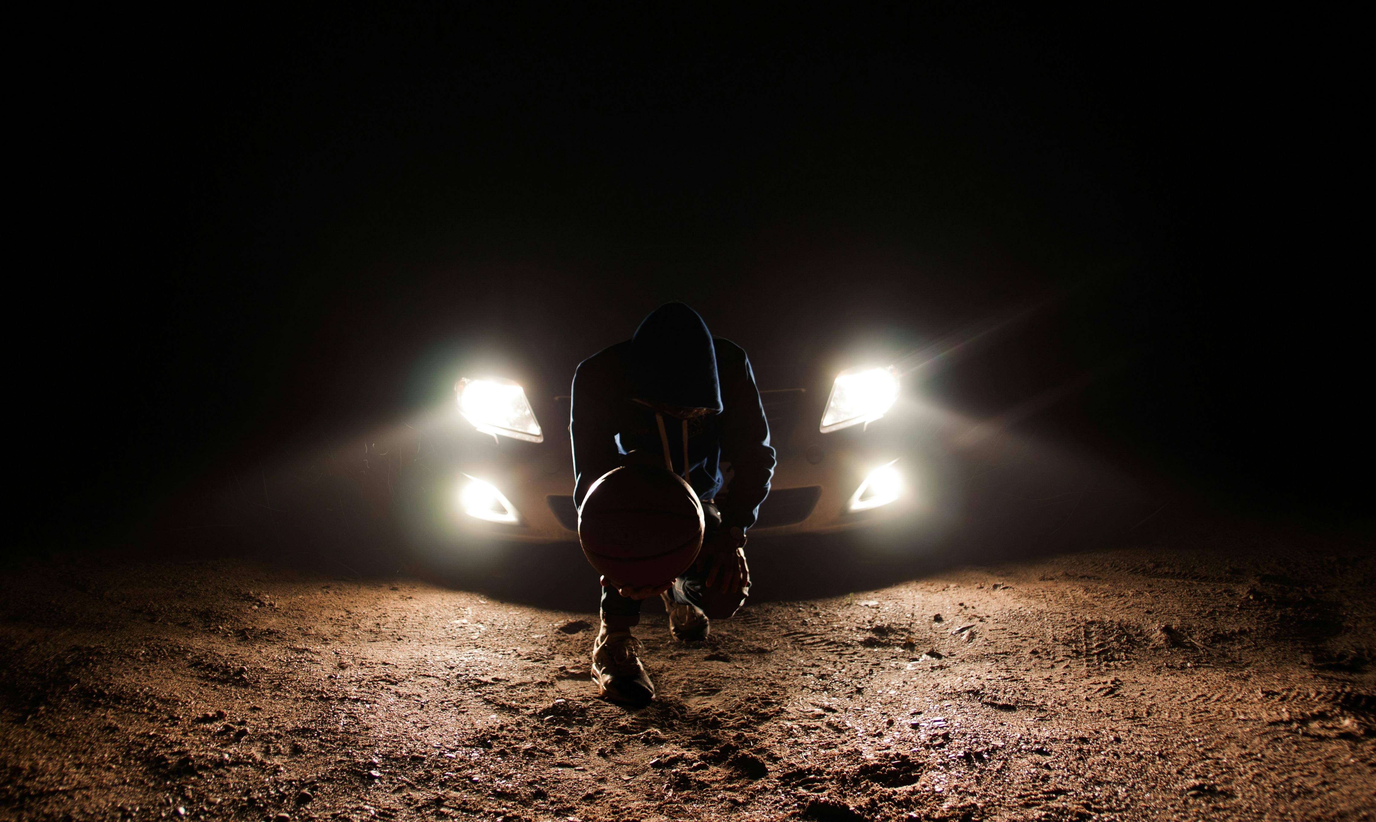 man against the vehicle headlight squatting holding ball