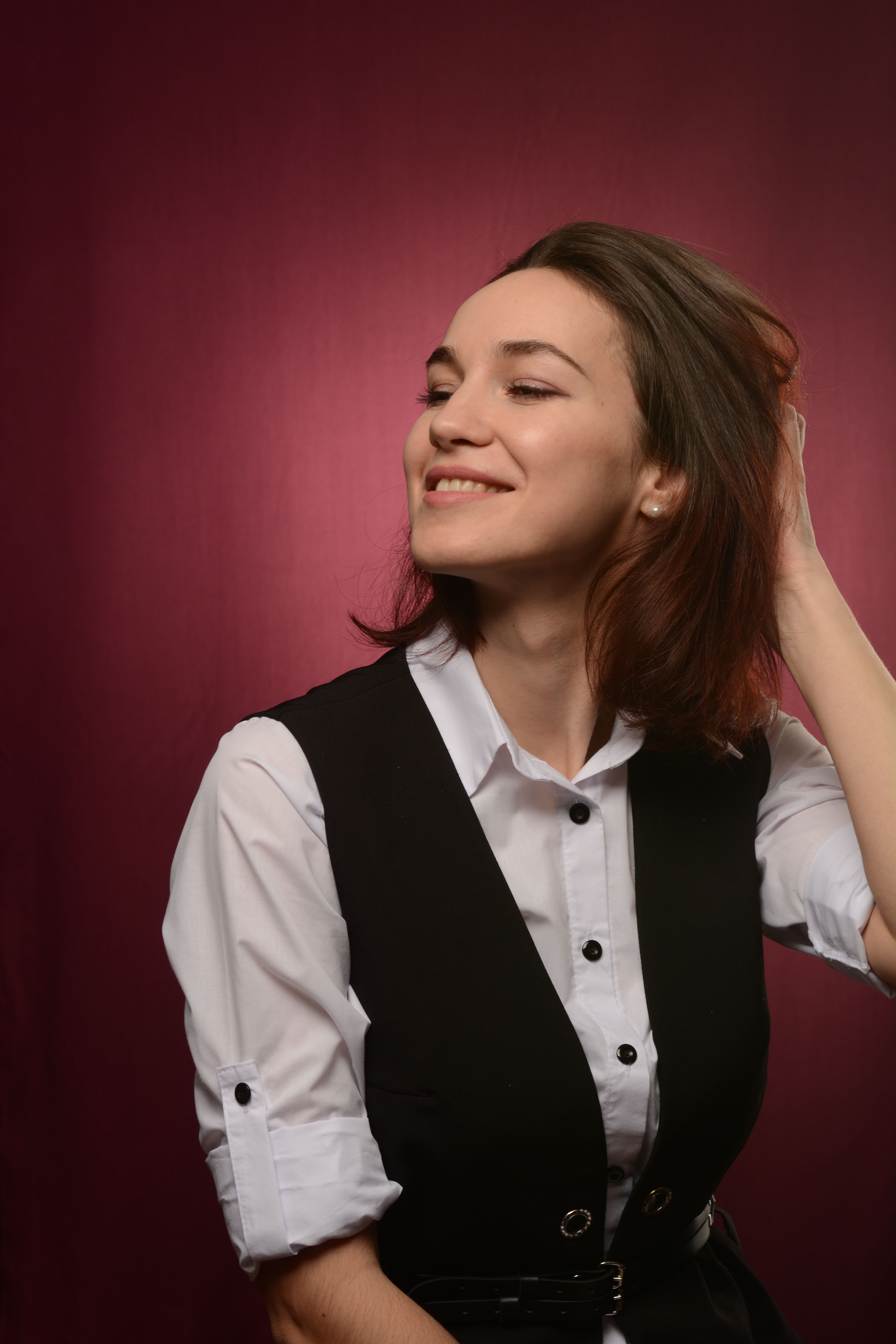 Elegant studio portrait of a woman smiling with hand on hair against a maroon background.