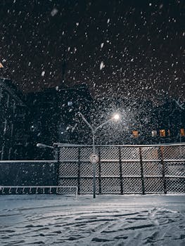 A snowy night scene in a city with streetlights illuminating falling snow and footprints.