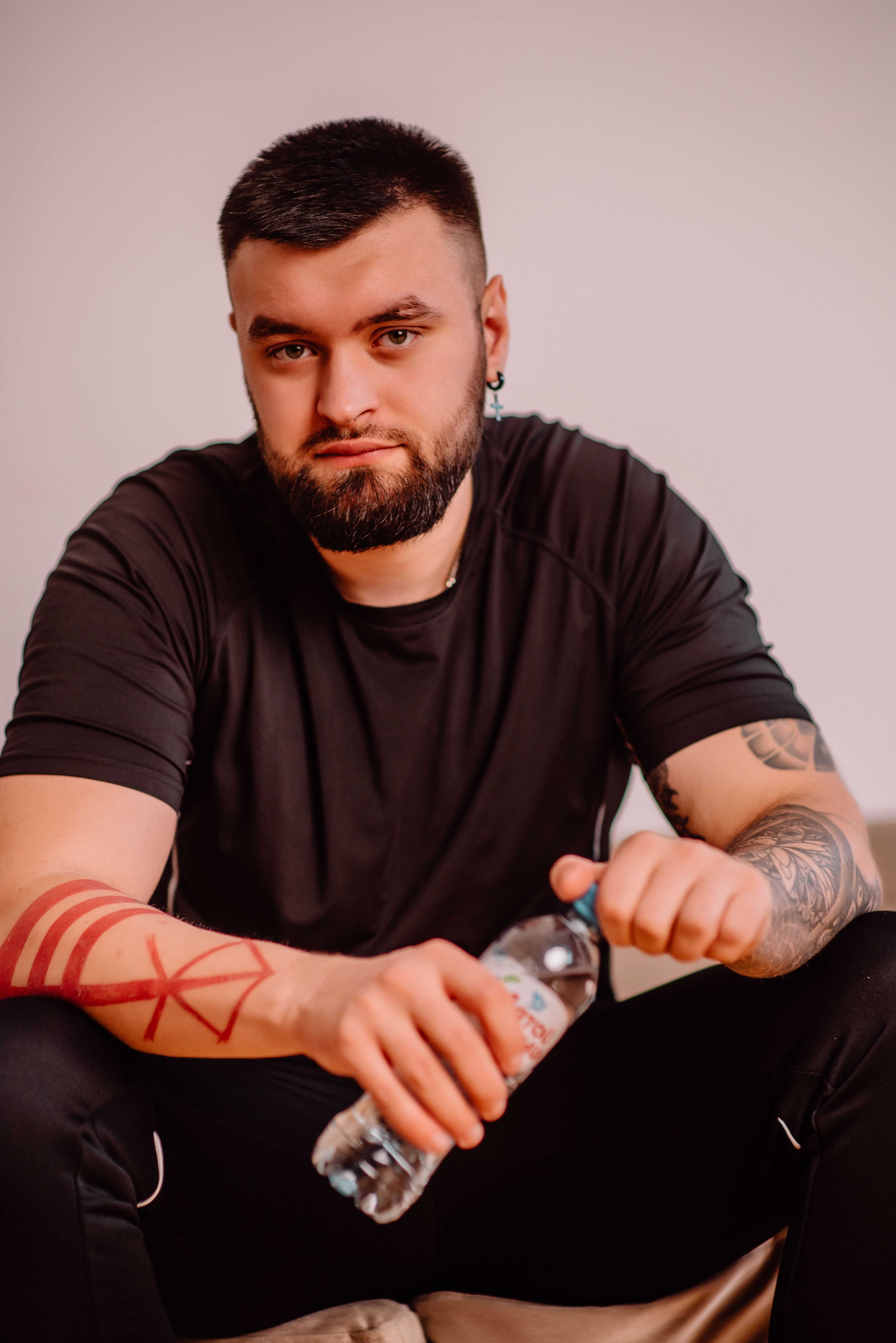 A bearded man with tattoos sits indoors holding a water bottle, wearing a black shirt.