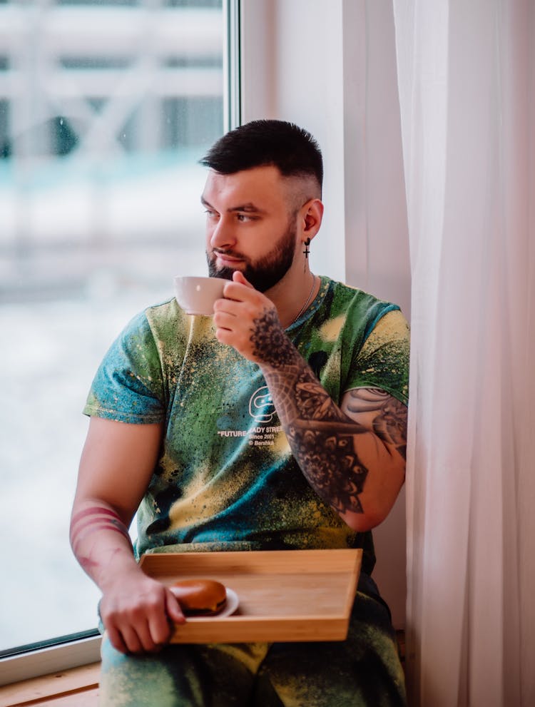 A Man Sitting Near The Window While Holding A Cup Of Coffee And A Wooden Tray