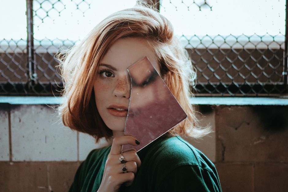 Como a Despersonalização Pode Impactar sua Autoestima Diária 2 Artistic portrait of a redhead woman holding a mirror, showcasing her freckles and unique expression.
