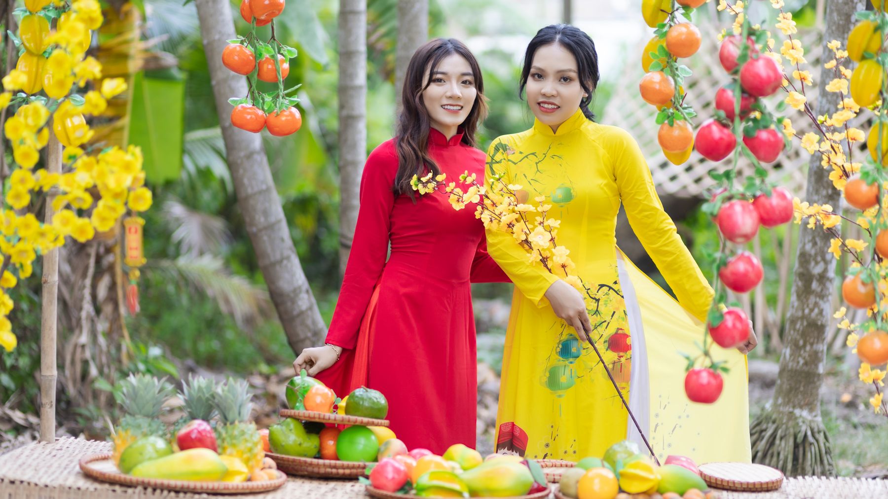 Hai người phụ nữ Việt Nam trong tà áo dài rực rỡ giữa khung cảnh trưng bày trái cây đầy màu sắc.