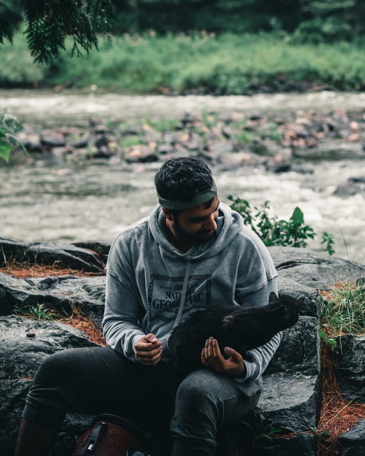 A Man In Hoodie Sweater Sitting While Holding A Black Cat