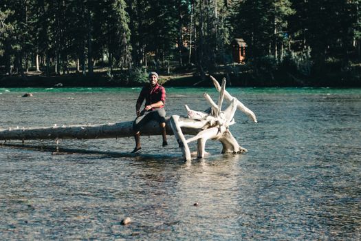A man sits on a fallen tree in a scenic Canadian river landscape, enjoying nature.