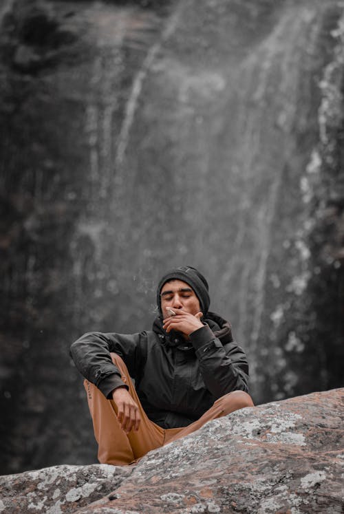 Man Crouching on a Stone with a Cigarette and Black Rock in Background