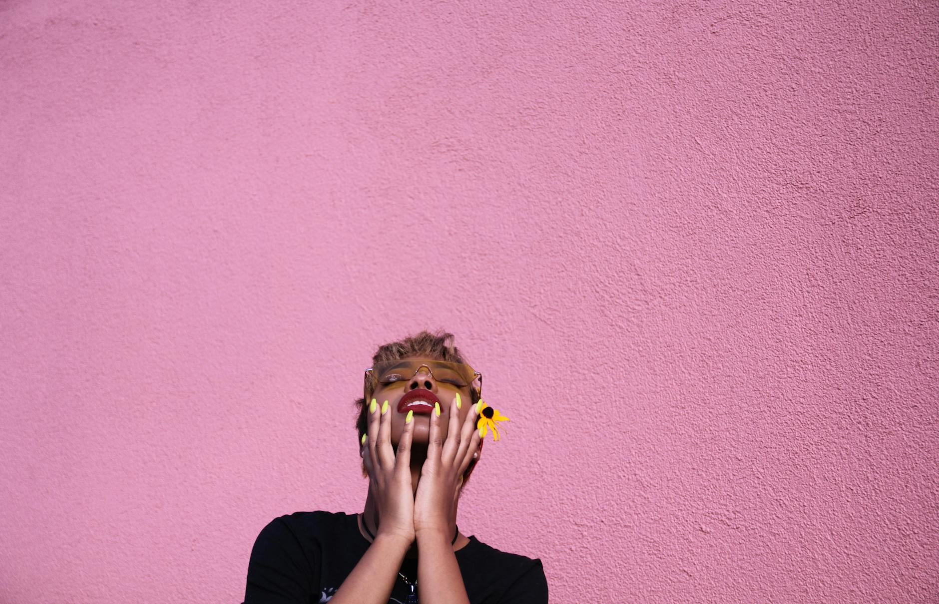 Stylish young African American female in casual wear and sunglasses with closed eyes touching face while throwing head back on pink background