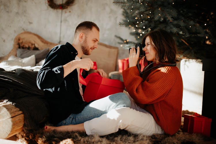 A Woman In Red Sweater Taking Picture Of A Man Opening A Present