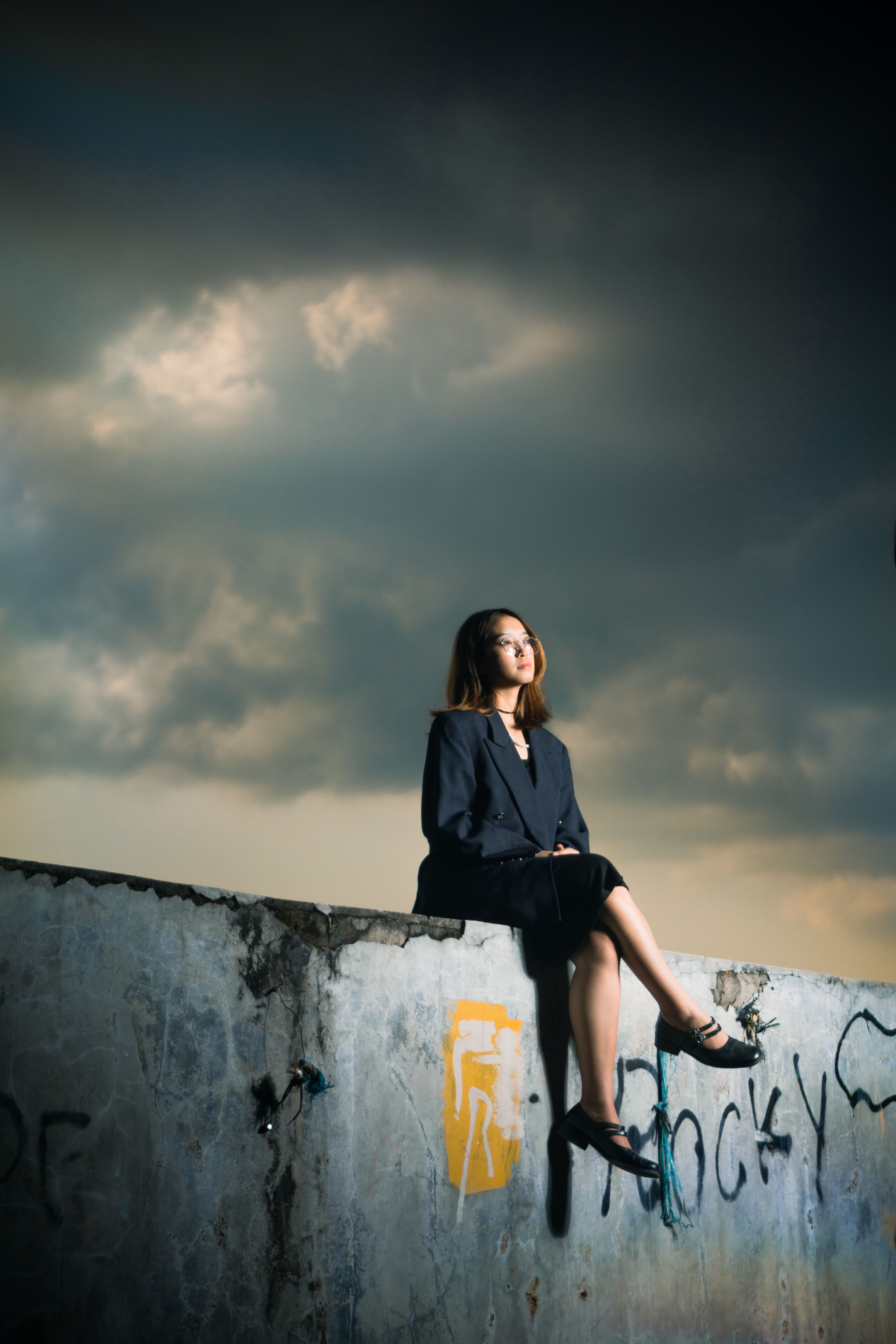 A stylish young woman sits on a graffiti-covered concrete wall under a dramatic cloudy sky in Bandung, Indonesia.