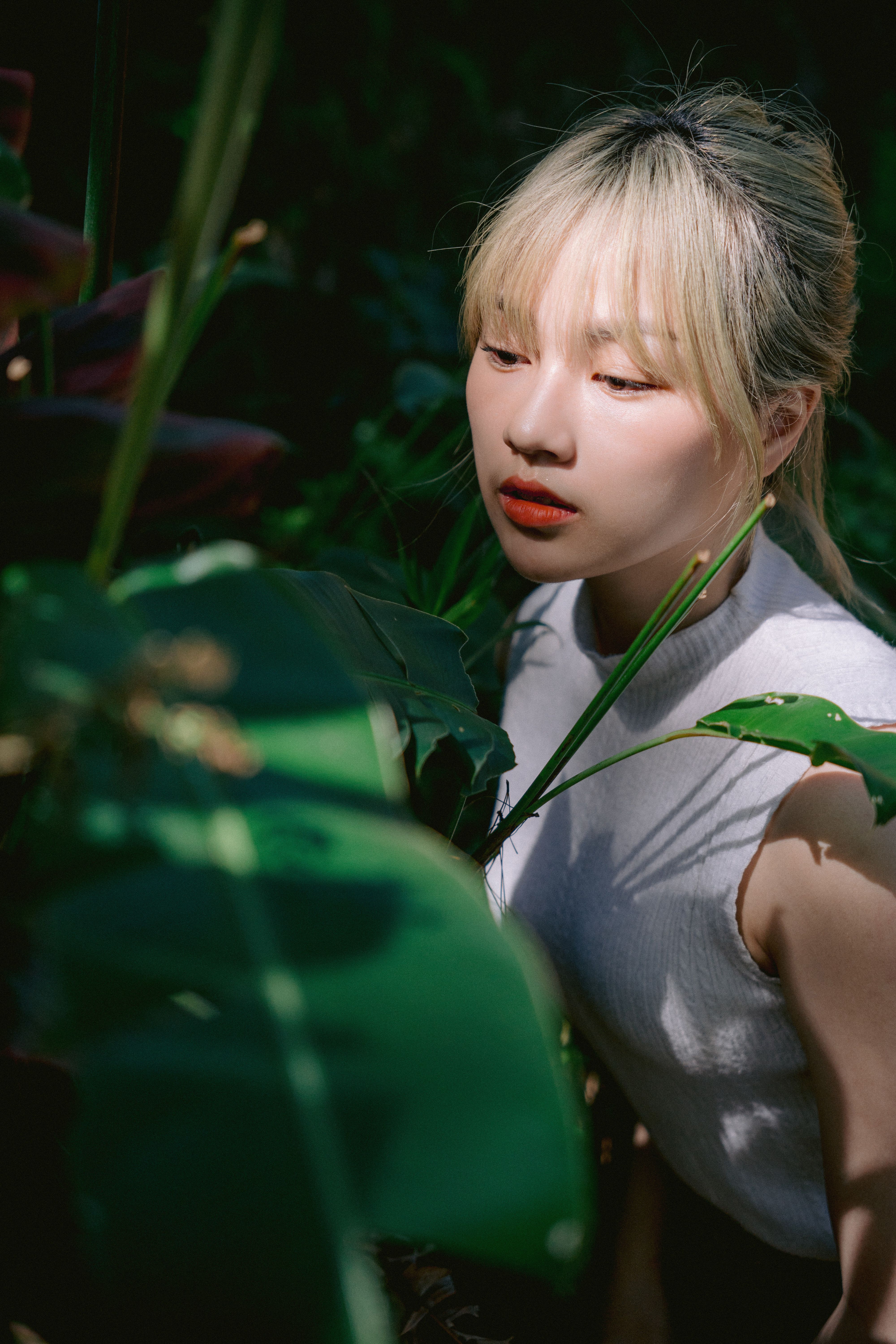 A young Asian woman stands surrounded by lush leaves in natural sunlight.